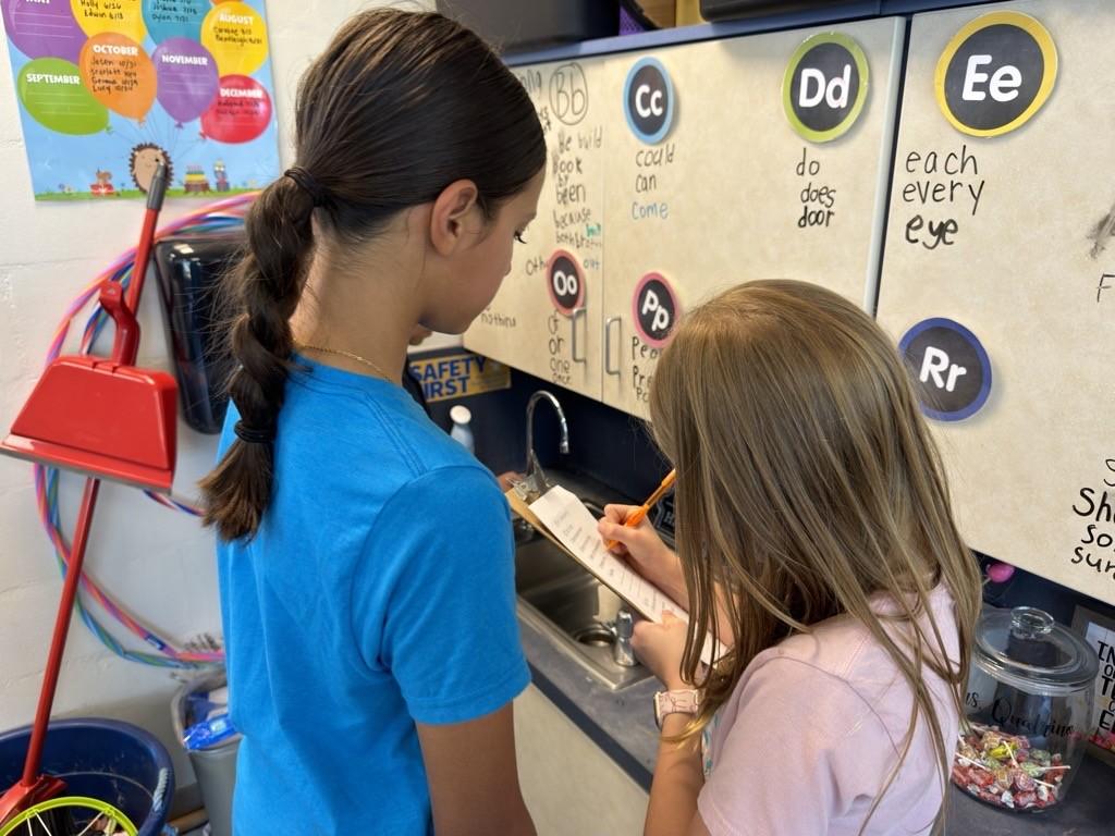 Two young students stand at a classroom sink area, where one girl in a blue shirt watches as another girl in a pink shirt writes on a clipboard near cabinets decorated with alphabet circles.
